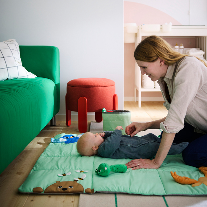 Smiling mother playing with her baby on a colourful quilt on the floor of a bright and cosy living room.