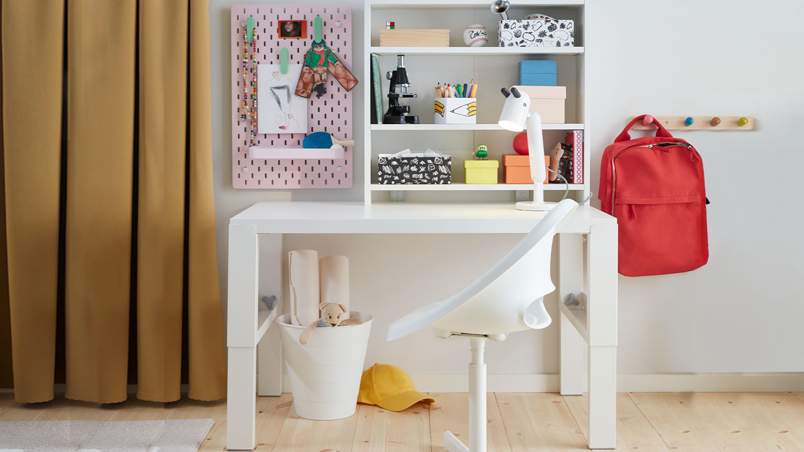 The white PÅHL desk placed in a children’s room with a white desk chair, a shelf holding colourful boxes and a pink pegboard