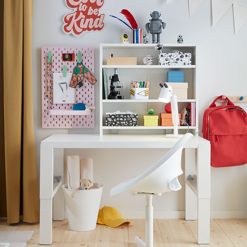 The white PÅHL desk placed in a children’s room with a white desk chair, a shelf holding colourful boxes and a pink pegboard