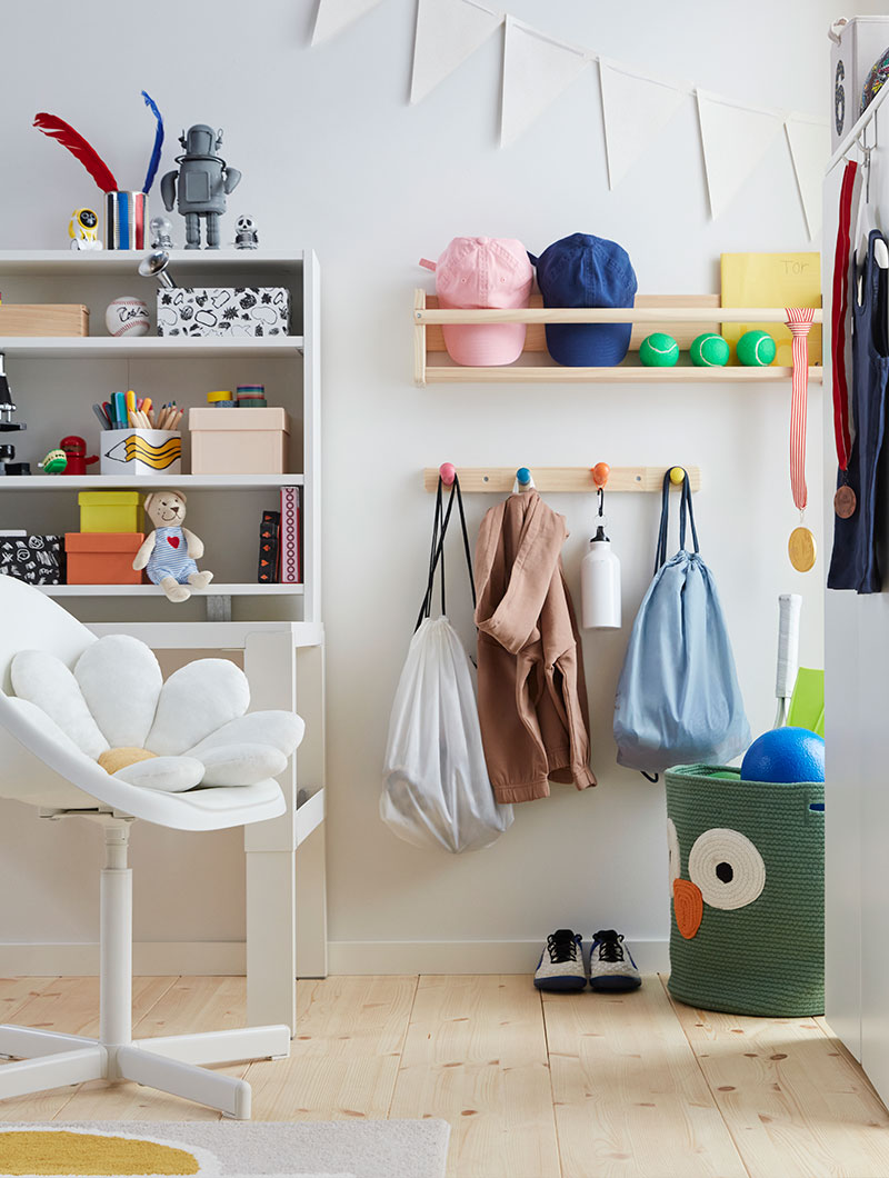 A children’s room with a FLISAT knob rack with four colourful knobs holding activity bags and clothes underneath a wall shelf