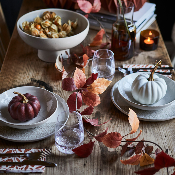 Mesa rústica de madera decorada para una cena de otoño con platos de cerámica, calabazas decorativas en blanco y burdeos, vasos de agua, cubiertos y hojas secas en tonos rojizos y marrones, con una vela encendida al fondo.