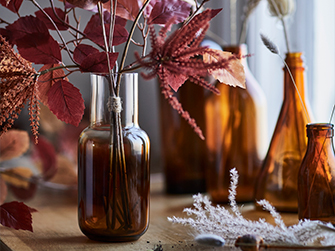 Primer plano de un jarrón de cristal ámbar con ramas decorativas de hojas rojizas, acompañado de otras botellas y jarrones de cristal ámbar difuminados al fondo, creando un ambiente cálido y otoñal.