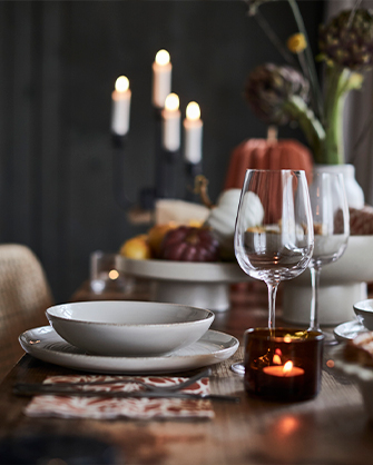 Detalle de una mesa de otoño con un plato de cerámica texturizada, una copa de vino vacía, un posavasos estampado y una vela encendida en un portavelas de cristal oscuro, con calabazas decorativas y un candelabro al fondo.