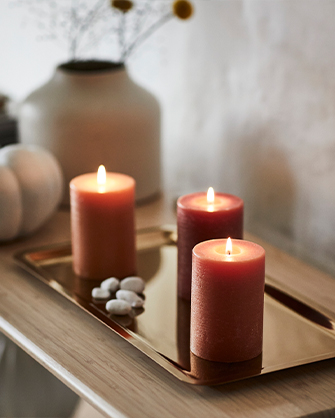Tres velas cilíndricas encendidas en tonos naranja y burdeos, colocadas sobre una bandeja dorada brillante, con pequeños guijarros blancos decorativos, creando un ambiente acogedor y cálido.