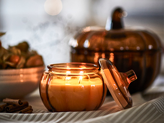 Vela de cera encendida dentro de un recipiente de cristal ahumado con tapa a juego, cerca de palitos de canela y un cuenco con physalis, todo con un suave difuminado en el fondo que sugiere un ambiente íntimo.
