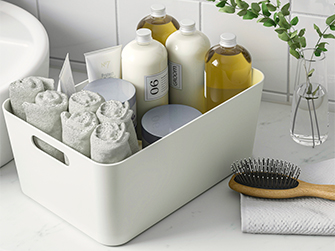 Caja de almacenamiento blanca en un baño, llena de productos de higiene personal y toallas enrolladas, facilitando la organización del espacio.