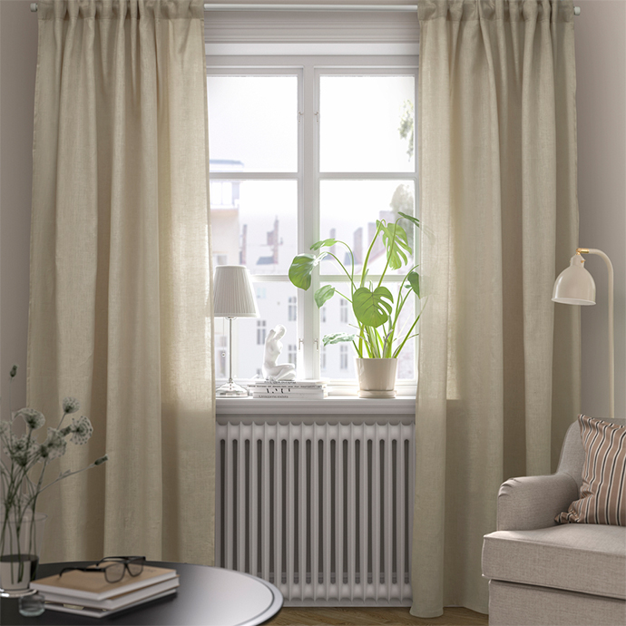 Ventana con cortinas largas de color beige claro y una planta de Monstera en el alféizar. Un radiador debajo de la ventana y una zona de estar con un sillón y una mesa baja.