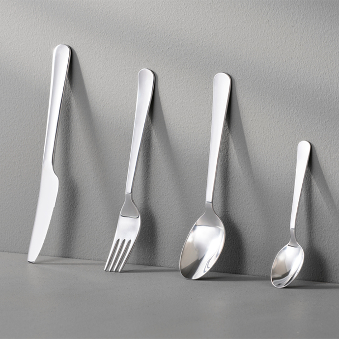 Stainless steel cutlery set with a matte finish, consisting of a knife, a fork, a spoon, and a teaspoon, leaning vertically against a light gray wall.