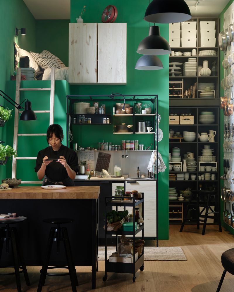 A person at a kitchen island photographs food on a plate. An ÄSPINGE kitchenette is placed by a green wall in the background.