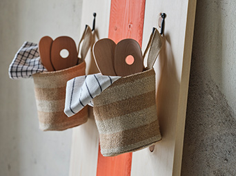 Two MÄVINN jute storage baskets filled with wooden utensils and tea towels hang by their loops on hooks from the wall.