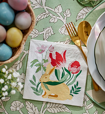 Un primer plano de una servilleta de papel TJÄRLEK con estampado de conejitos de Pascua, junto a un plato UPPLAGA sobre un mantel verde SMÖRFISK.