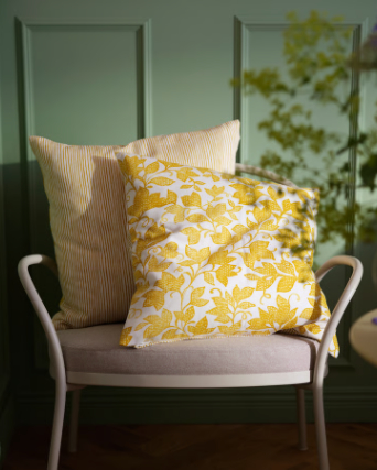 A yellow and white FJUNKLOCKA cushion cover rests on a light armchair, surrounded by green walls and soft light.
