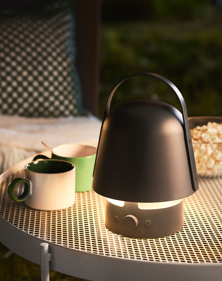 A garden setting features a blue VAPPEBY bluetooth speaker lamp with a rounded design, standing on a table with drinks.