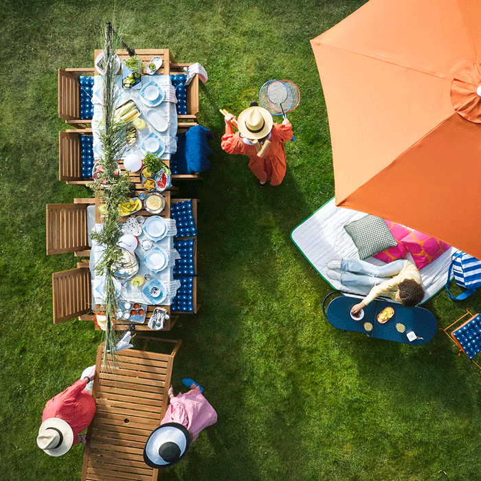 Vista aérea de una fiesta de verano en el jardín con una larga mesa de madera, una sombrilla naranja y personas disfrutando en el césped.