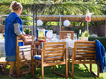 Comedor exterior con mesa y sillas de madera con cojines azules, decorado con un dosel de hojas frescas y farolillos de papel blancos.