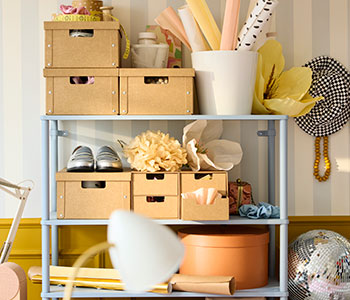 A bright wall with a light grey-blue SÅGMÄSTARE shelving unit, displaying organised boxes and decorative items on shelves.