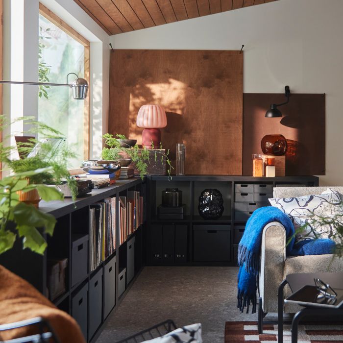 Una sala de estar cuenta con una estantería KALLAX de color marrón oscuro con libros y objetos decorativos junto a un sofá gris.
