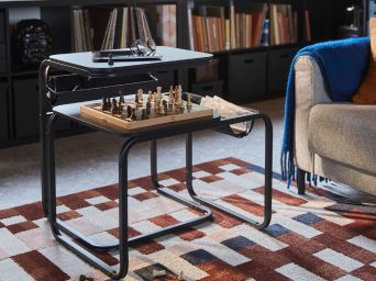 Una sala de estar con un juego de ajedrez sobre una mesa auxiliar OLSERÖD junto al sofá y sobre una alfombra MARKBLÅMME de pelo corto estampada.