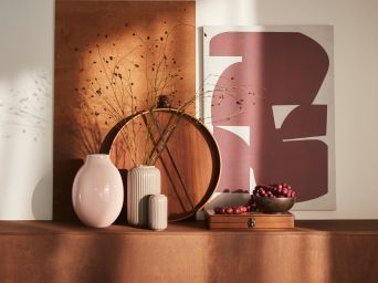 Un jarrón BLODBJÖRK de color rosa pálido se coloca sobre una superficie de color tostado junto con más jarrones, objetos decorativos y un cuadro PJÄTTERYD.