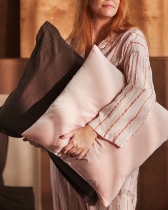 Una persona sostiene una almohada con una funda HAMPFLOCKEL de color marrón oscuro y un cojín con una funda SANELA de color rosa pálido.