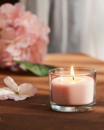 Primer plano de una vela perfumada LOTSFÅGEL rosa pálido en un recipiente de cristal colocada sobre una mesa con flores rosas al fondo.