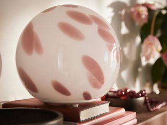 Primer plano de una lámpara de mesa FADO rosa de cristal que descansa sobre unos libros apilados junto a un cuenco marrón y una planta.