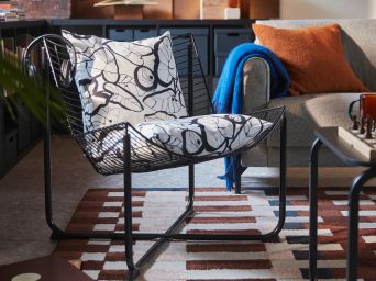 Una sala de estar cuenta con un sillón SKÅLBODA negro con dos fundas de cojín PELARKÖRSBÄR, una mesa de centro de madera y una alfombra suave.