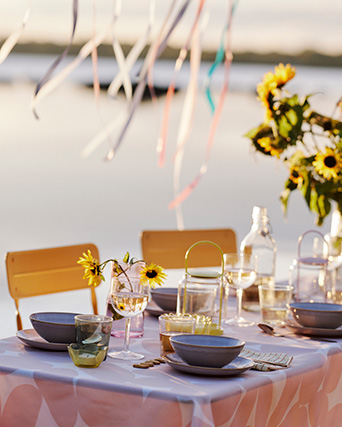 Festive lakeside table with sunflowers, dinnerware, and colorful ribbons under soft light.