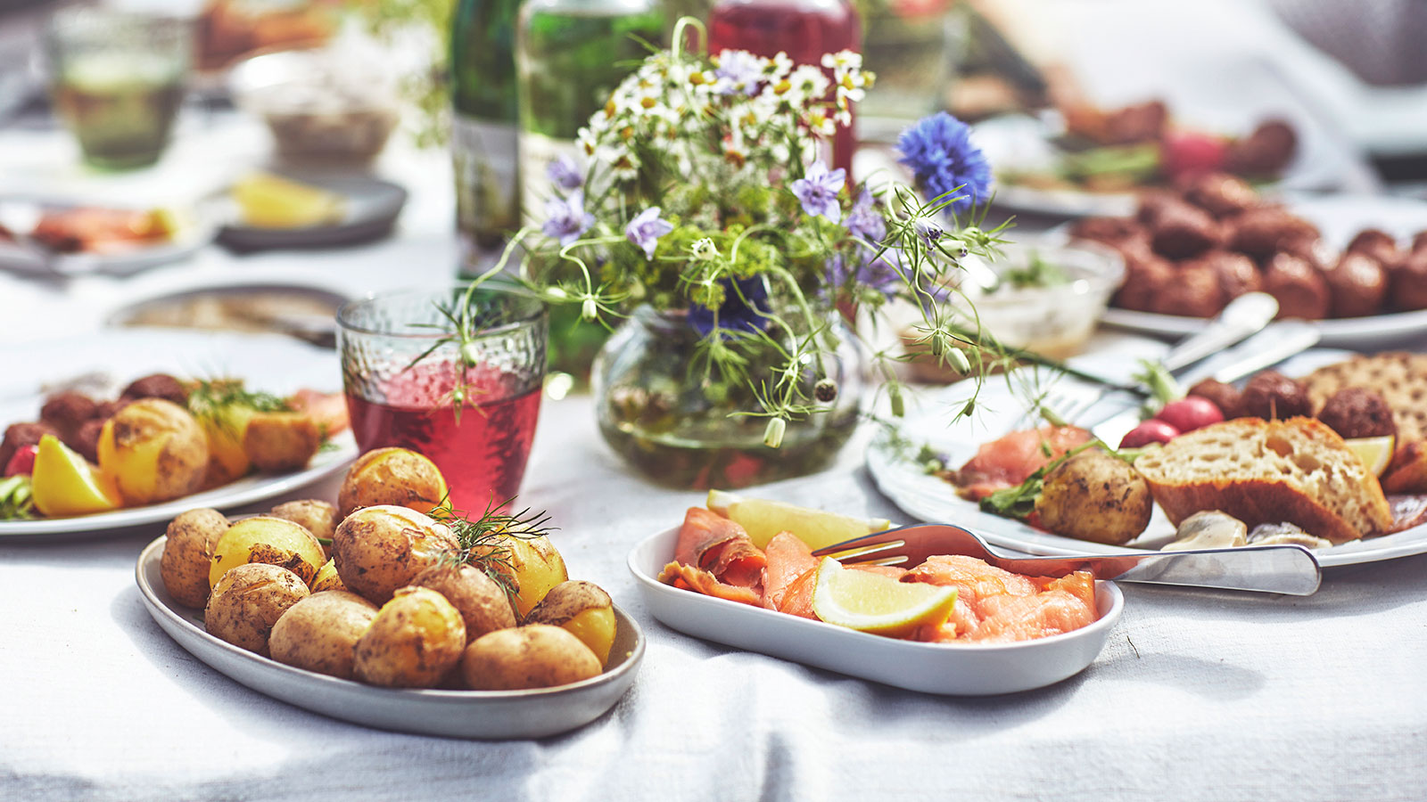  Outdoor table setting with roasted potatoes, salmon, meatballs, and a vase of wildflowers, with a sparkling water view in the background. 