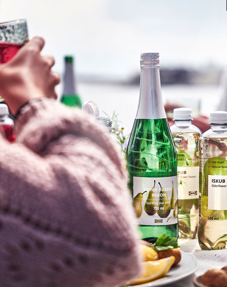 Table set with bottles of sparkling pear drink and elderflower water, alongside shared plates of food during an outdoor gathering by the water.