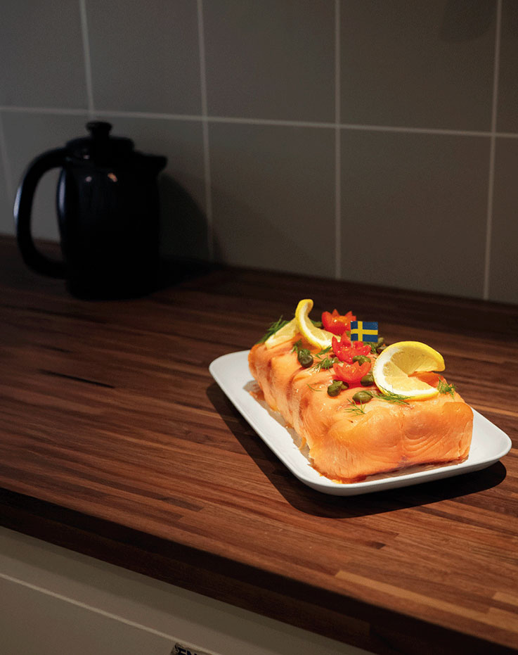  Salmon dish garnished with lemon slices and fresh dill, served on a white rectangular plate on a dark wooden countertop near a black kettle.