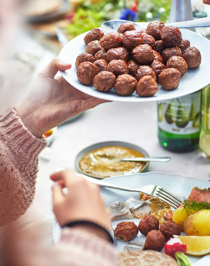  Close-up of a person in a pink knit sweater holding a white plate of meatballs over a table filled with various dishes and potatoes