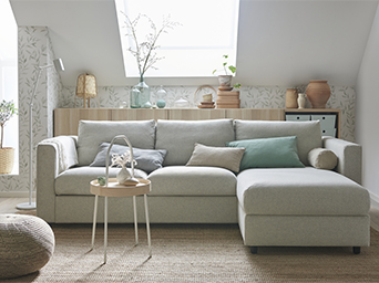 Sofá seccional gris claro en una sala luminosa con decoración de estilo natural, papel tapiz botánico y una mesa auxiliar de madera.