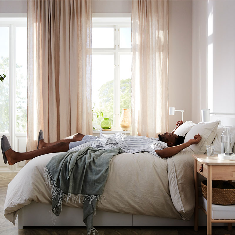 Image of a brightly lit bedroom with beige curtains in side view, with a man lying down as if he just 
