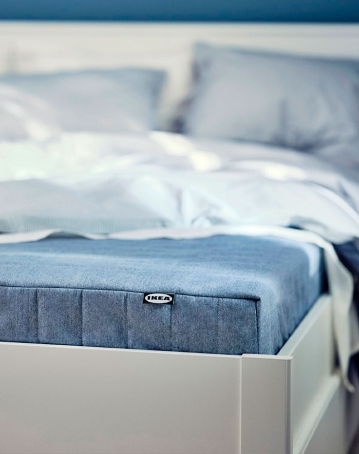 Corner of a white modern style bed, with the mattress visible in blue and a white quilt on top.