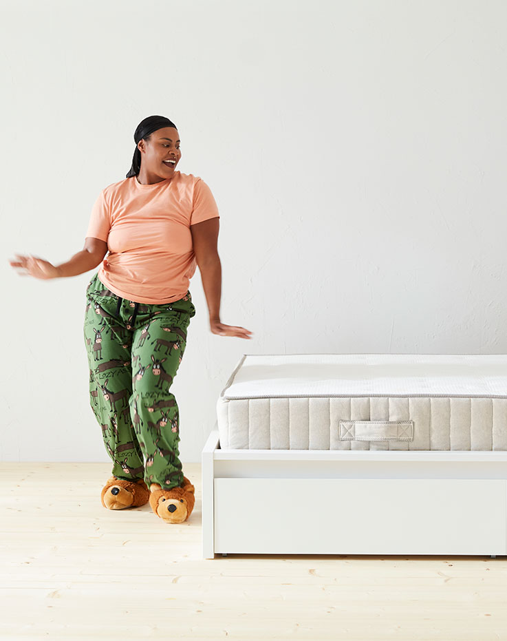 Woman standing in a dynamic pose in front of a bed with a white mattress visible, the shot is fully illuuminated and white.