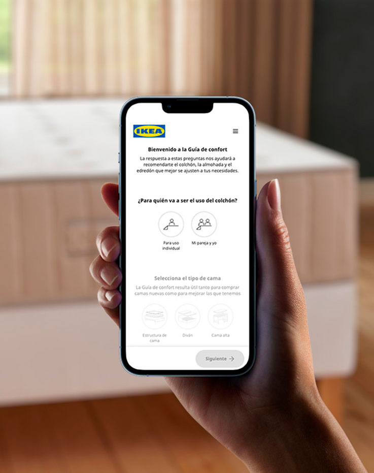 Hand holding a cell phone, on the screen you can see a mattress guide app, in the background out of focus you can see a large beige mattress.