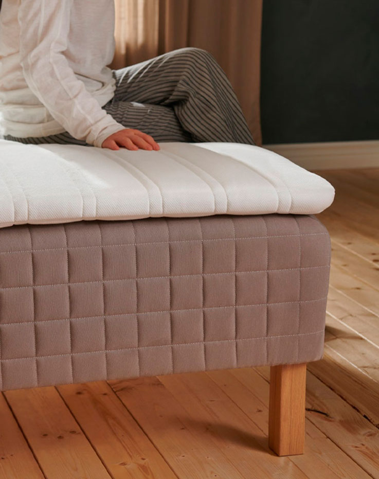 a person sitting on a bed, the mattress is visible, it looks big and on top of it there is a small mattress, the image only shows the corner of the bed and only part of the body of the seated person.