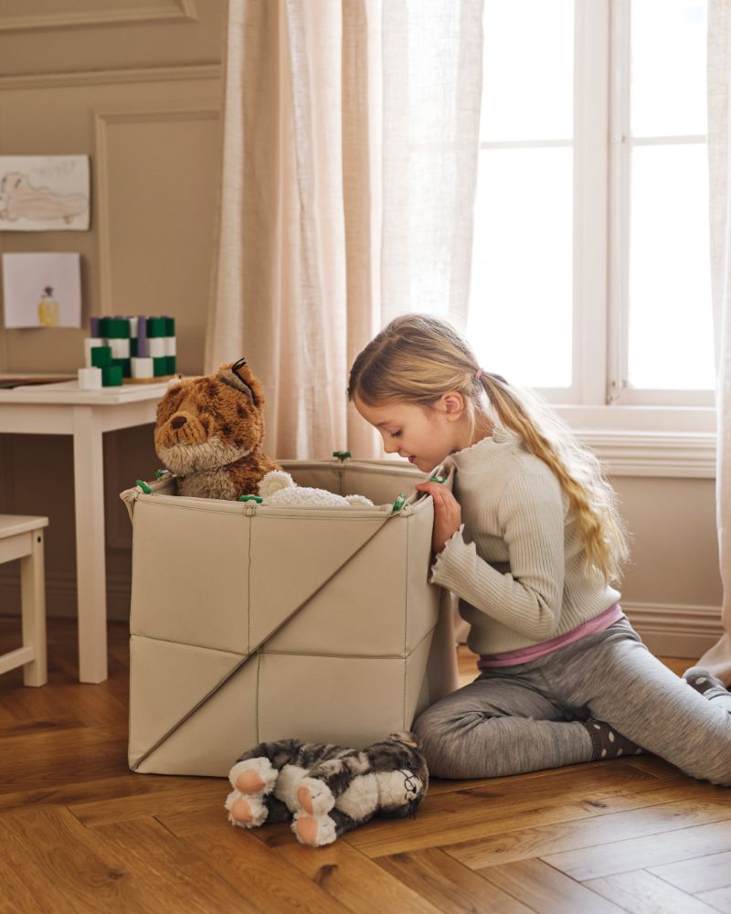 A child looks inside a GREJSIMOJS mat/storage box containing toys on the floor beside a white SUNDVIK children’s table.