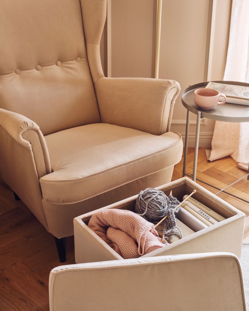 A deep beige STRANDMON footstool with its lid off and a STRANDMON wing chair stand in a living room near a tray table.