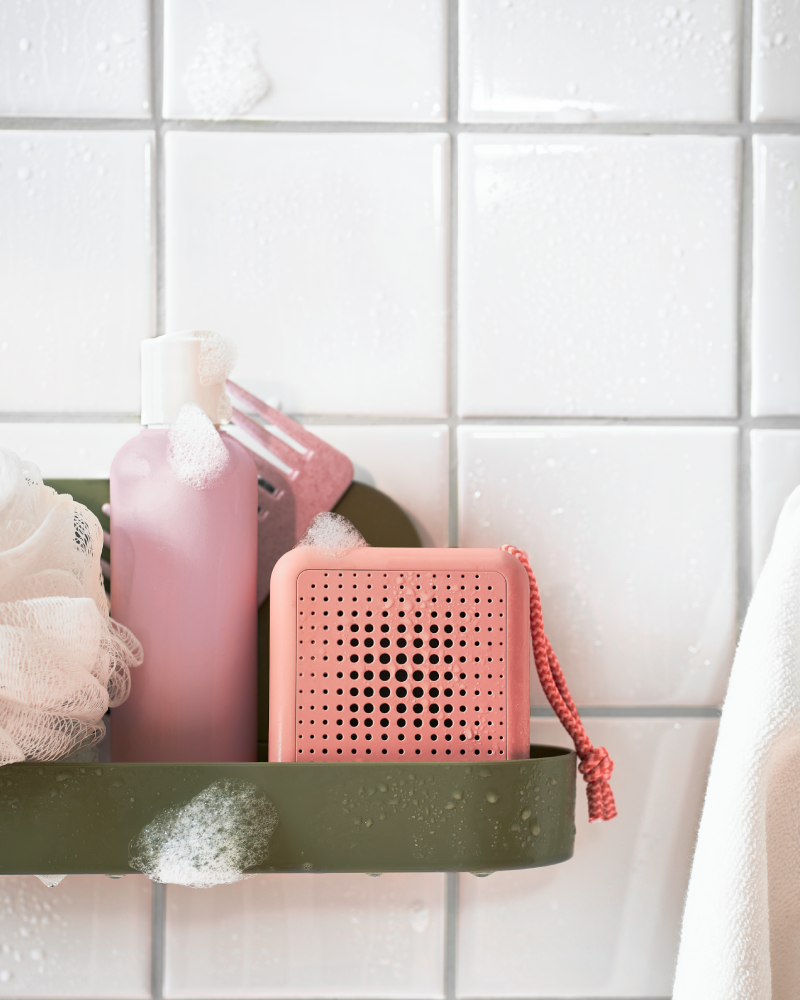 Wall shelf with suction cup with a greyish-green suction cup, featuring a beige sponge and a pink waterproof portable speaker.