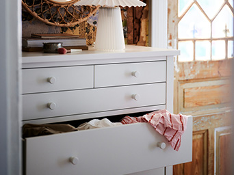 White dresser with open drawers, a pleated lamp on top, and a rustic wooden door in the background.