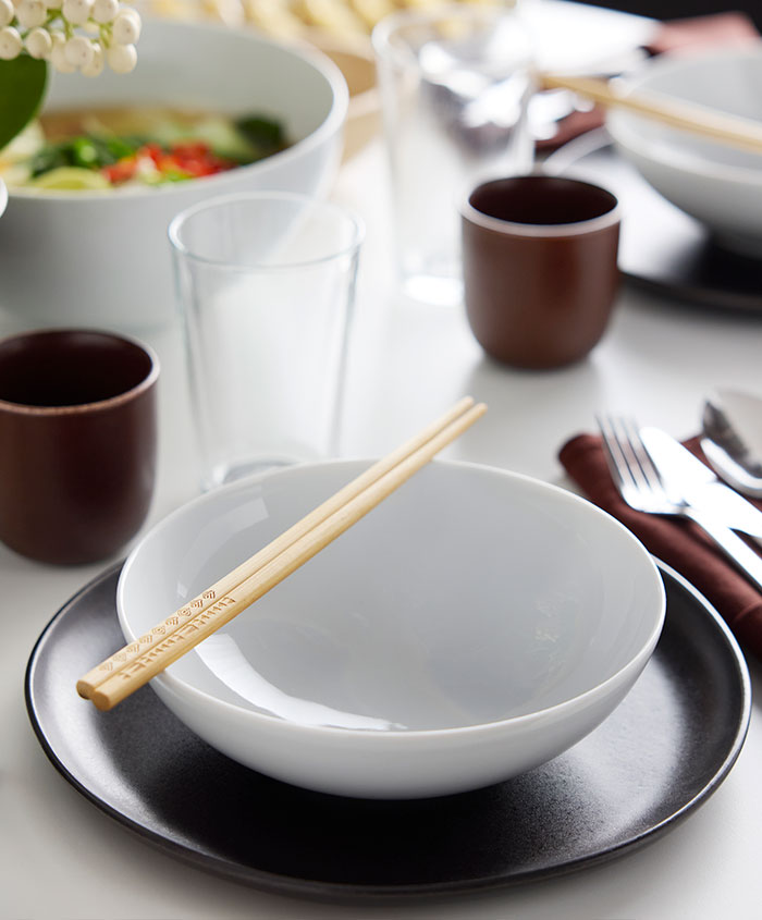 Two chopsticks lie on top of an IKEA 365+ bowl which stands on a black STOCKHOLM 2025 plate near an IKEA 365+ glass.