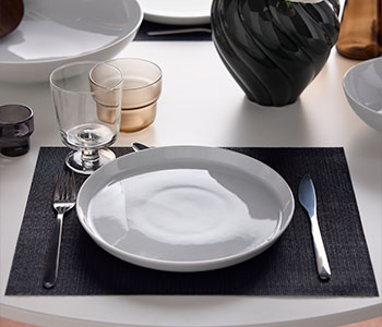 Un servicio de mesa compuesto por un plato de la colección IKEA 365+, una copa de la colección IKEA 365+, cubiertos de la colección PRAKTBOTIA y un vaso OLIVTRAST de color marrón claro.