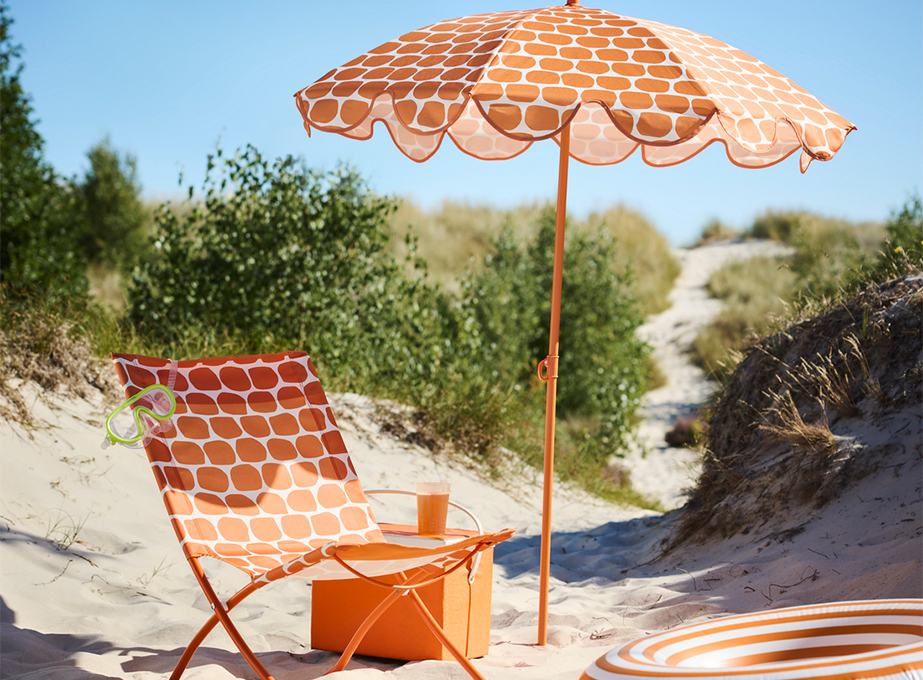 An orange and white patterned beach chair and matching umbrella set up on a sandy dune path, accompanied by an orange cooler box, a drink, and a snorkel mask.