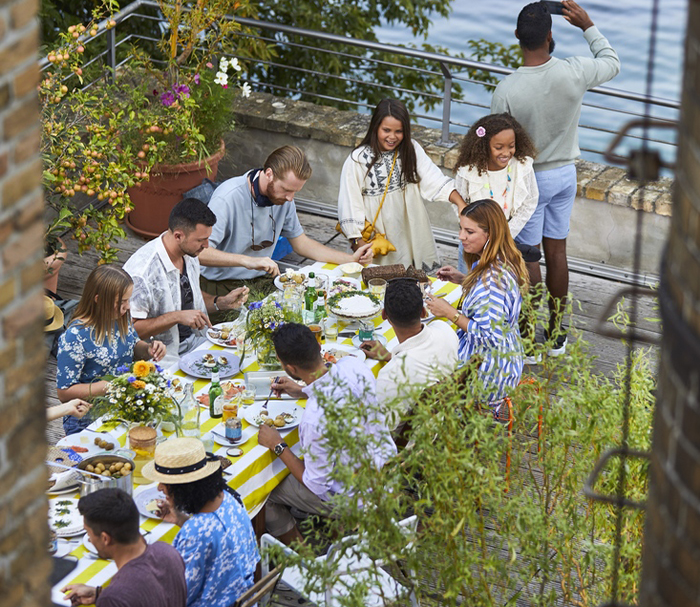 A high-angle view of a large group of family and friends gathered around a long table with a yellow and white striped tablecloth, enjoying an outdoor meal on a rooftop terrace.