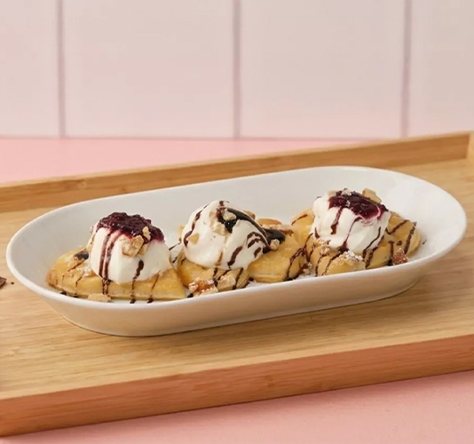 Three heart-shaped waffles topped with ice cream, chocolate syrup, berry jam, and crushed nuts in an oval white dish.