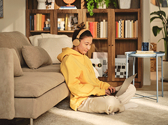 A person sitting on a rug, leaning against a beige sofa while using a laptop with headphones in front of a wooden bookcase.