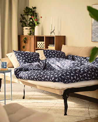 Detail of an unfolded sofa bed with a blue and white patterned duvet, pillows, and a wooden shelf unit in the background.