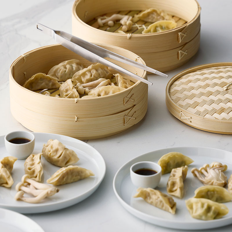A dining setting with a bamboo KLOCKREN steamer filled with dumplings and two small bowls of dipping sauce.